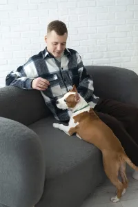 A young man in a plaid shirt sits comfortably on a grey sofa with a brown and white dog resting beside him