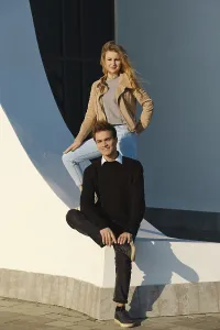 A young man sits on a low wall with his leg bent while a woman stands above him