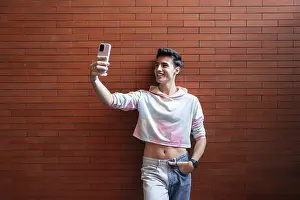 A young man smiles while taking a selfie with his smartphone against a vibrant red brick wall