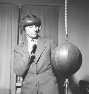 A young man in a suit stands with his guard up. He intends to hit a cue ball that is attached to the floor and ceiling, also called a floor-ceiling ball. With no gloves and only head protection during his training, he is ill-equipped for the task