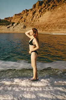 A young redhead woman stands on salt formations in the Dead Sea by the shores of Jordan