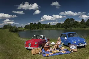 Category Oldtimer Prints: Two young woman having a picnic next to two vintage cars at the lake in sunny weather