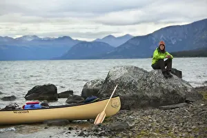 Young woman sitting on a rock, canoe and wooden paddle on the shore