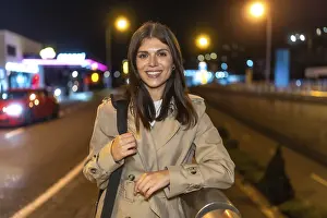Young woman smiling while standing in a trench coat on an urban street at night